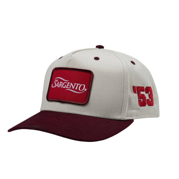 Angled side view of "The Heritage Snapback", showing a red front patch, cream crown, and burgundy accents.