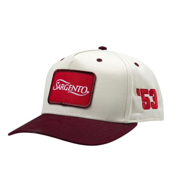 Angled side view of "The Heritage Snapback", showing a red front patch, cream crown, and burgundy accents.